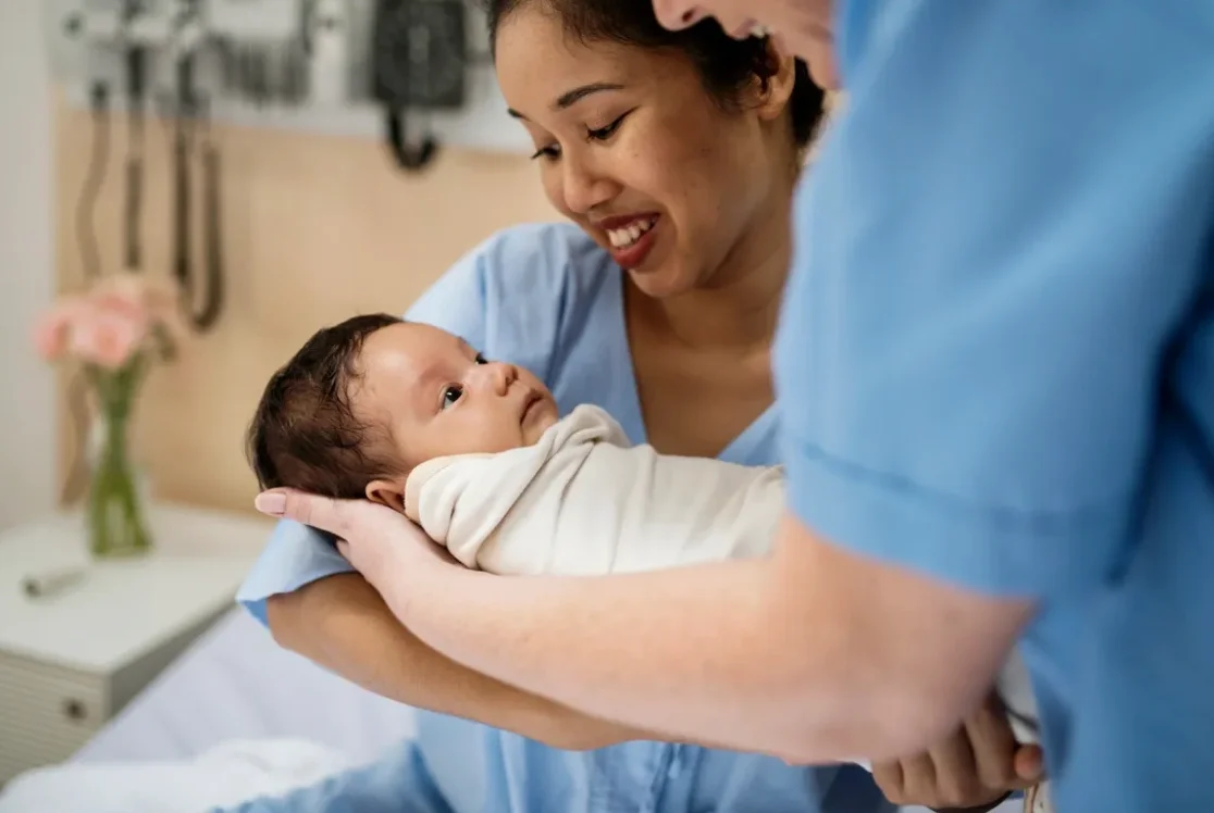 Newborn held by a woman in a hospital exam room as a clinician in scrubs stands beside them